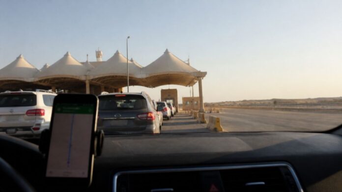 Absher Travel for Visitors: cars queue at a Saudi land border crossing while a phone on the dashboard shows a travel app